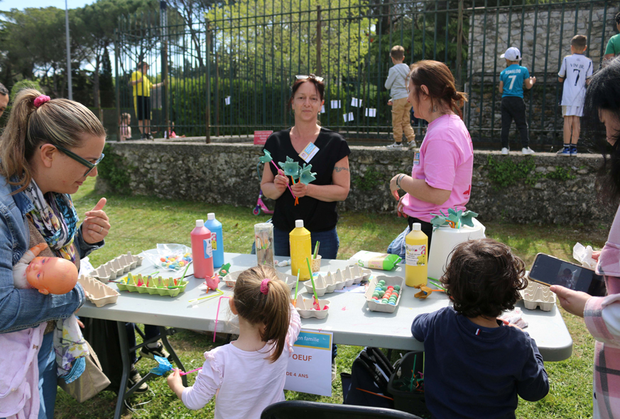 En extérieur, des adultes animent un atelier de création de fleurs avec des boites d'oeufs pour des enfants.