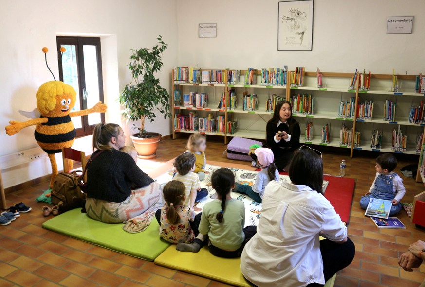 Dans une salle de médiathèque avec des livres sur les étagères, une adulte anime un atelier de lecture de contes devant un petit public d'enfants et de parents.