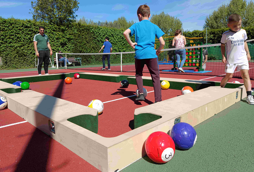 Sur un terrain de tennis extérieur, des enfants jouent à un jeu en forme de billard géant dans lequel les boules sont remplacées par des ballons de foot. Il s'agit de snookball.