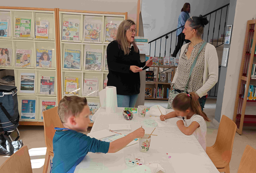 Dans une salle, deux adultes échangent tandis que deux enfants réalisent des dessins et de la peinture sur une table en participant à un atelier jeux créatifs.