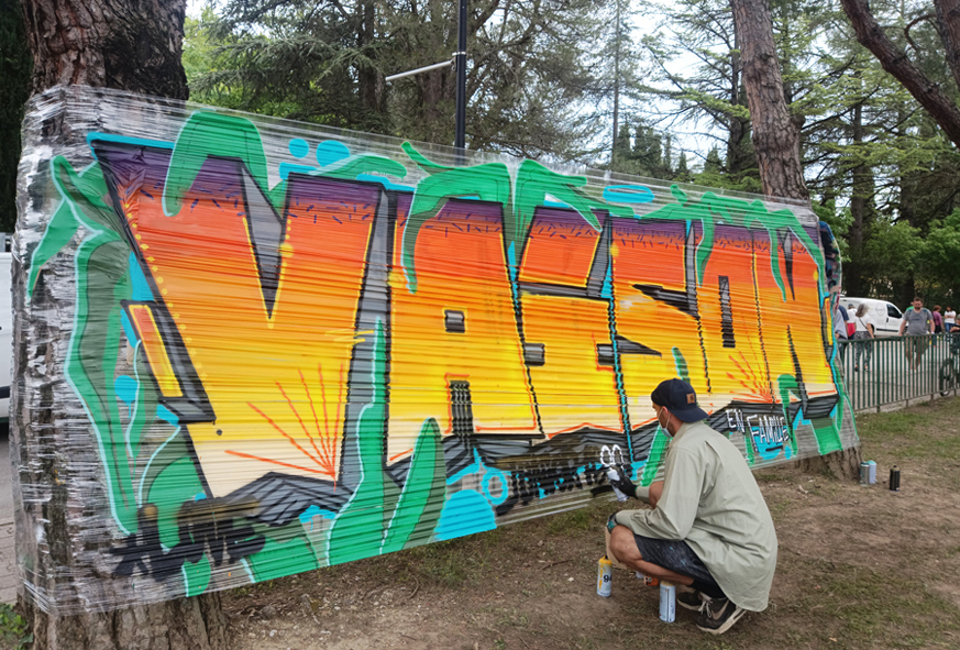 En extérieur, un graffeur peint une fresque avec le mot "VAISON" sur un film plastique tendu entre deux arbres.