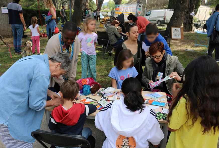 En extérieur, des enfants et adultes participent autour d'une table à un atelier créatif pour fabriquer une poupée tracas.