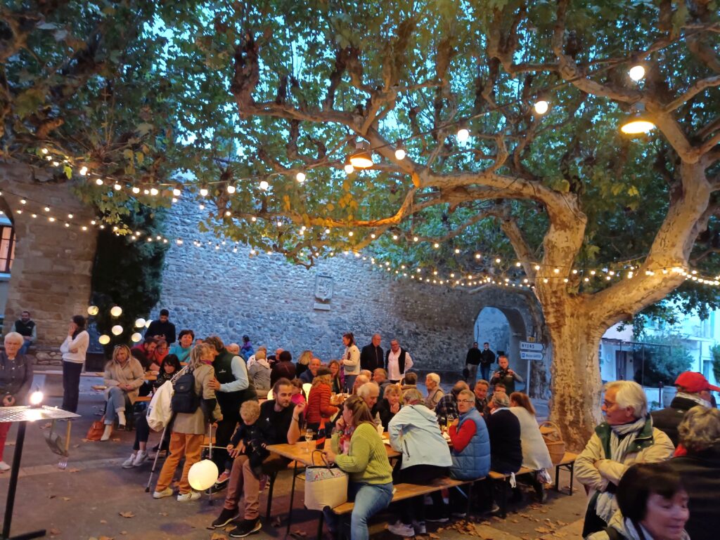 De nombreuses personnes attablées sur une place provençale à l'automne, sous des platanes et des guirlandes lumineuses.