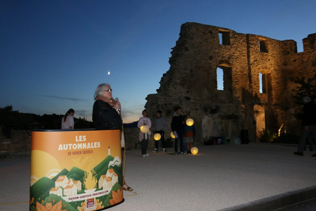 Une femme tient un micro sur la place de Faucon, à la tombée de la nuit. On voit les remparts de Faucon et une signalétique à l'effigie des Automnales.